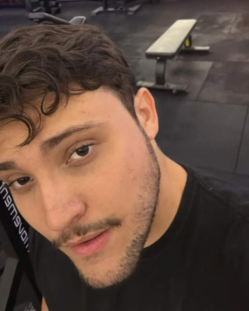 Selfie de Fillipe na academia, de camiseta preta, com bancos e aparelhos ao fundo